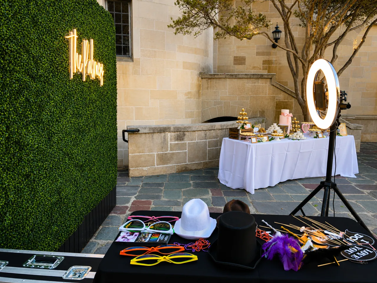 An open-air photo booth with a green hedge backdrop, neon sign and props table